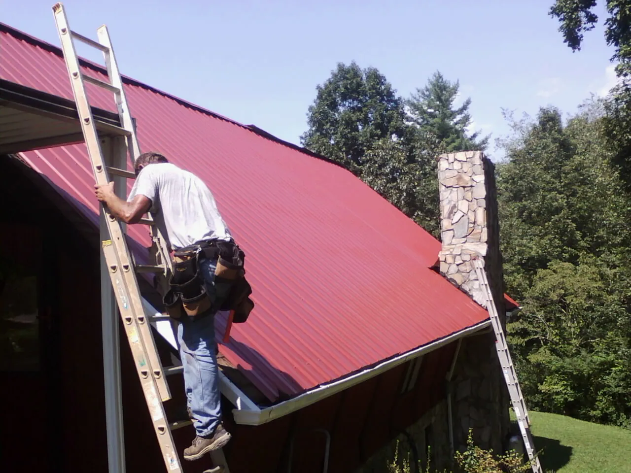 Roofer on a ladder beside a red metal roof and stone chimney — Soffit & Fascia Repair by Roofing Brothers in Hickman